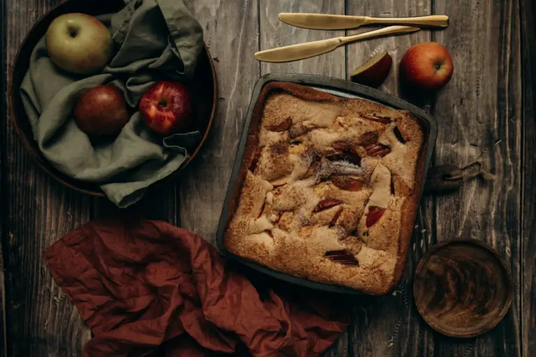 Ciasto z jabłkami upieczone w blaszce, obok świeże jabłka na drewnianym stole