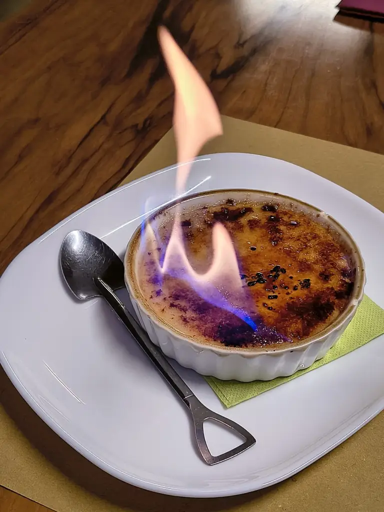 Crème brûlée w białej kokilce z płonącą karmelową skorupką, obok łyżeczka na białym talerzu